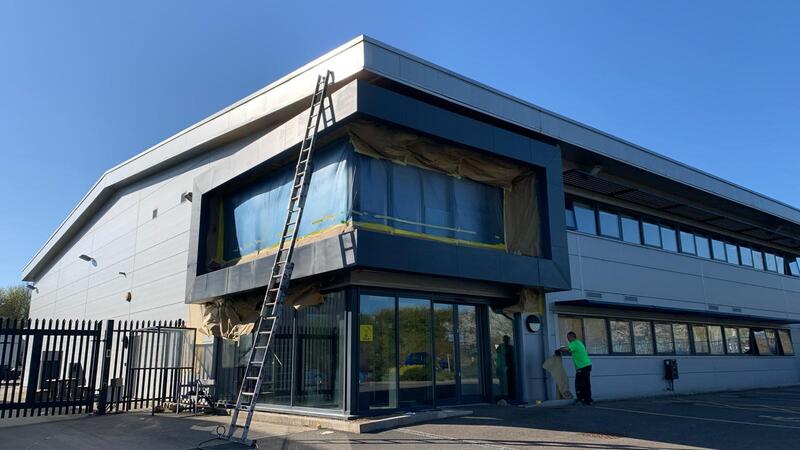 A worker on a ladder spray painting the dark grey upper facade of a commercial building with masking sheeting in place.