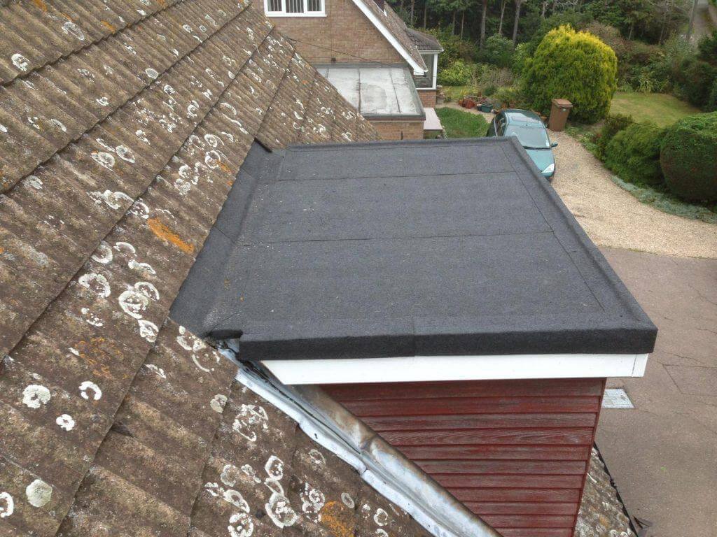 A small flat felt roof over a dormer extension, seen alongside an older lichen-covered tiled roof with a car parked below.