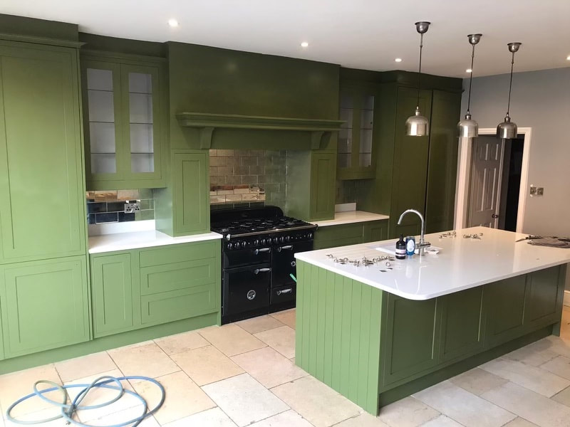 A kitchen with olive green painted cabinets, a white island, and a black range cooker beneath a tiled splashback.