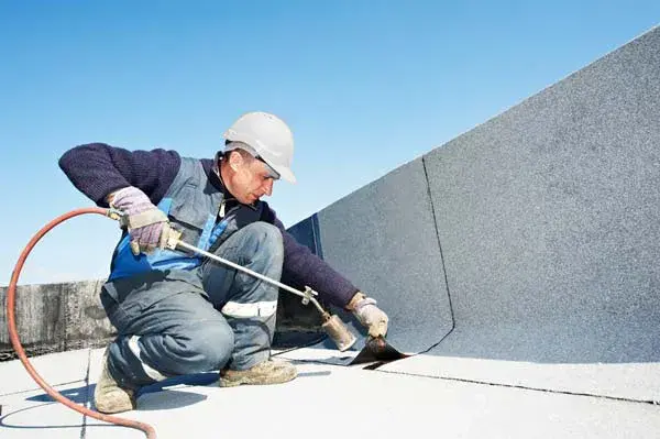 A roofer in a hard hat using a gas torch to heat-weld felt roofing on a flat roof edge.